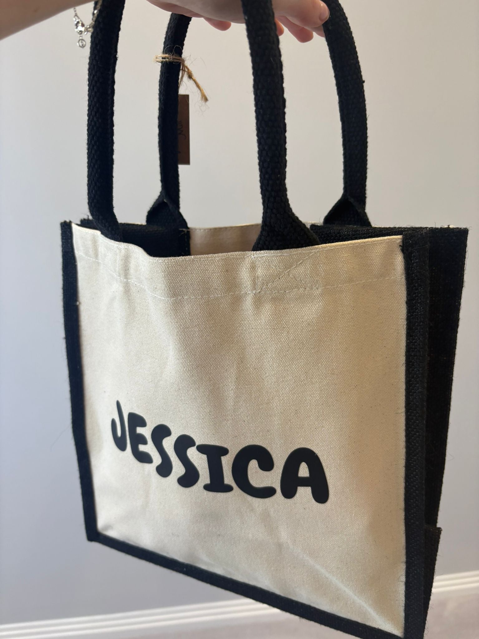 Beige personalised tote bag with 'JESSICA' printed on it, held by a hand against a neutral background.