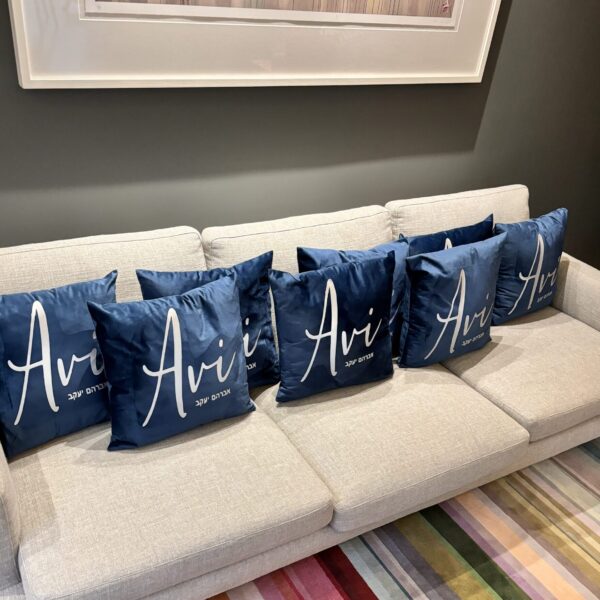 Beige sofa with personalised blue silk pillow cases featuring the word 'Avi' on a grey wall background.