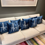 Beige sofa with personalised blue silk pillow cases featuring the word 'Avi' on a grey wall background.