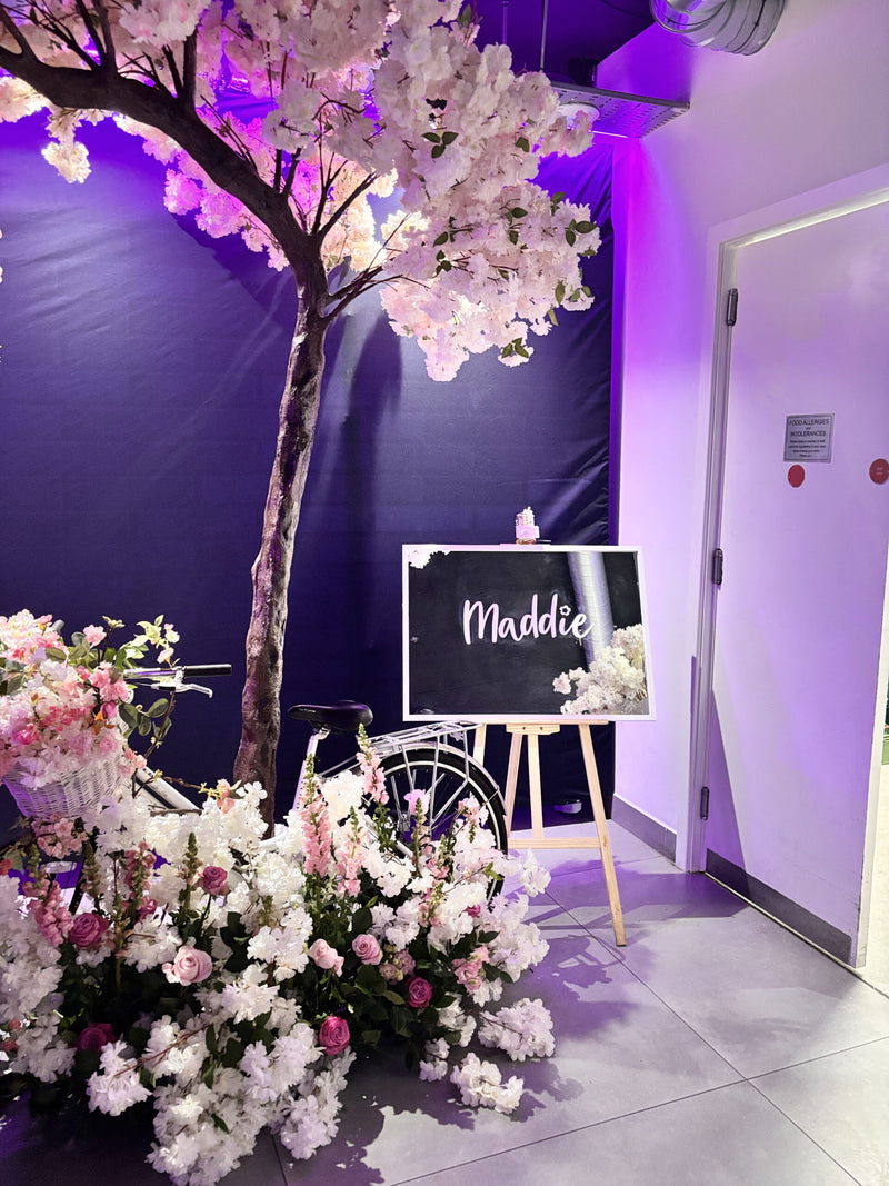Decorative floral arrangement with a tree and 'Maddie' mirror sign in a room with purple lighting.