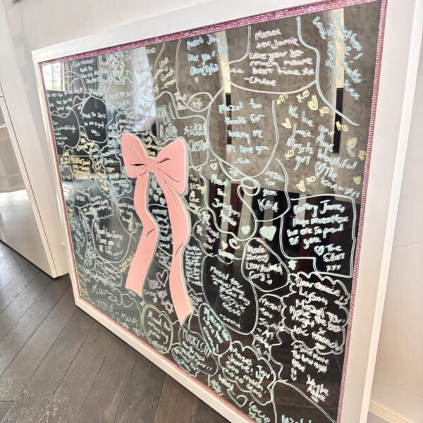 Decorative bespoke mirror with a pink bow and handwritten messages on a wooden floor.
