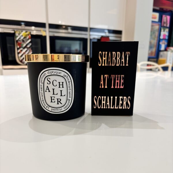 Black candle with gold lid and 'Schaller' branding next to a match box sleeve reading 'Shabbat at the Schallers' on a reflective surface.