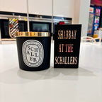 Black candle with gold lid and 'Schaller' branding next to a match box sleeve reading 'Shabbat at the Schallers' on a reflective surface.