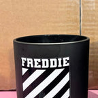 Black cylindrical candle with 'FREDDIE' printed on it against a cardboard background