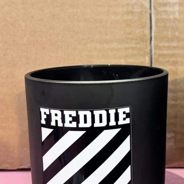 Black cylindrical candle with 'FREDDIE' printed on it against a cardboard background