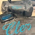 Black duffel bag and black washbag and black personalised pillow with 'Elo' text in blue on a wooden surface.