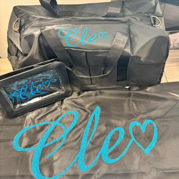 Black duffel bag and black washbag and black personalised pillow with 'Elo' text in blue on a wooden surface.