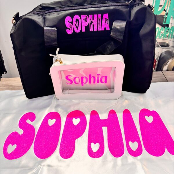 Black duffel bag and pink make up bag with 'Sophia' printed on them, placed on a white personalised pillow with 'Sophia' text.