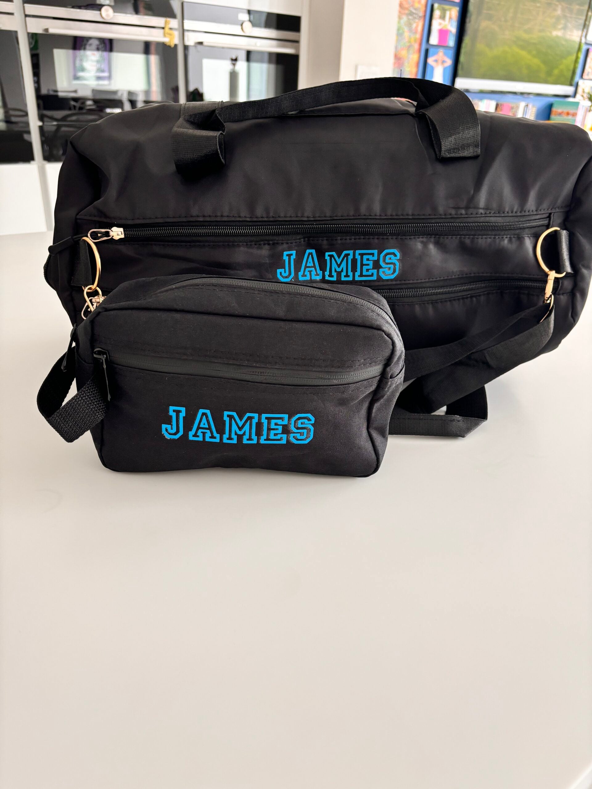 Black duffel bag and washbag and small pouch with 'JAMES' printed on them, placed on a light surface.