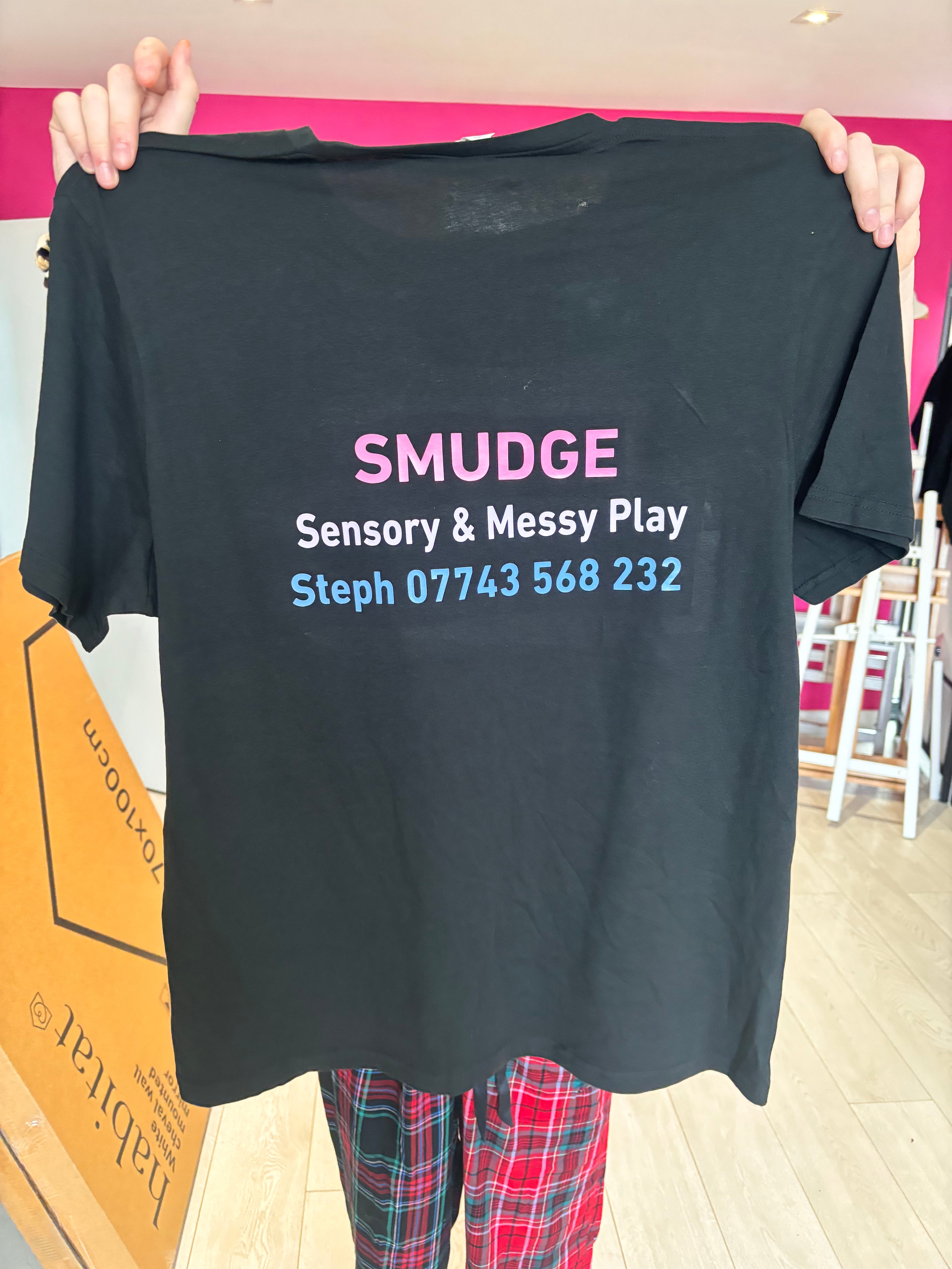 Black event t-shirt with 'SMUDGE Sensory & Messy Play' text held by a person.