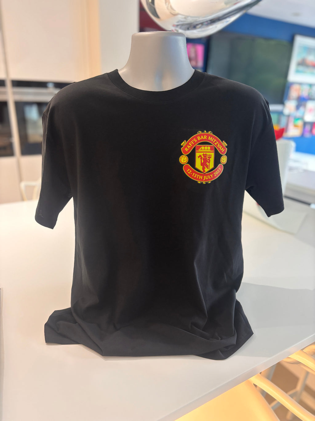 Black event t-shirt with a red and yellow Manchester United football club emblem on a mannequin in a store setting.