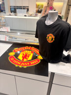 Black event t-shirt with a red and yellow Manchester United football logo on a mannequin in a store setting.