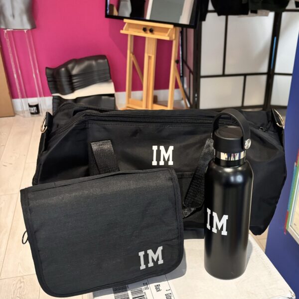 Black washbags and water bottle with 'IM' initials on a surface with a colourful background.