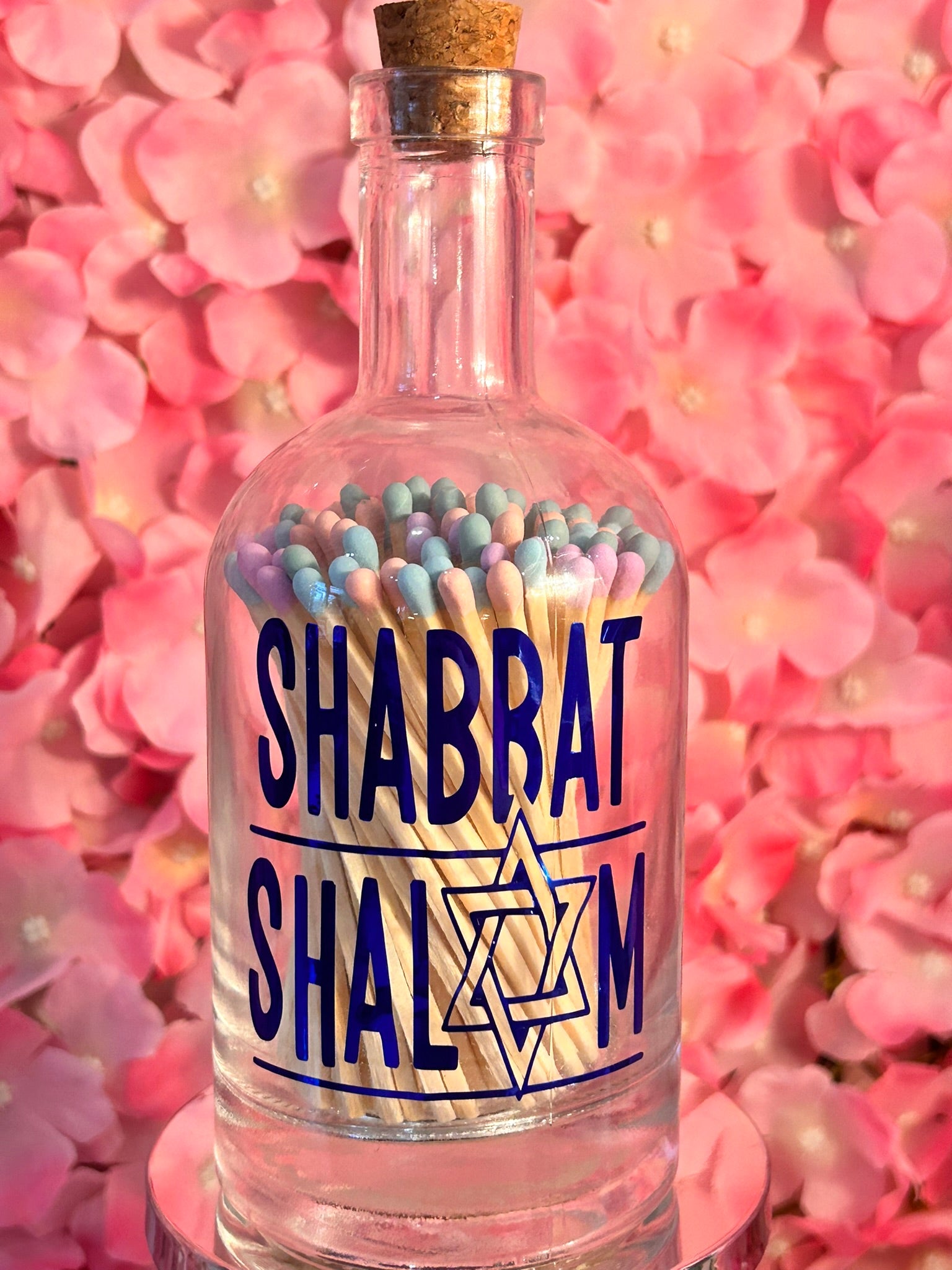 Clear glass bottle matches with 'Shabbat Shalom' label and matchsticks against a pink floral background