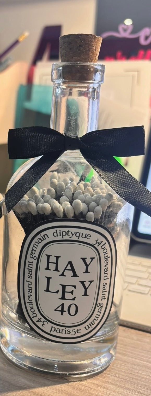 Clear glass bottle matches with black ribbon and Diptyque-style label on a wooden surface