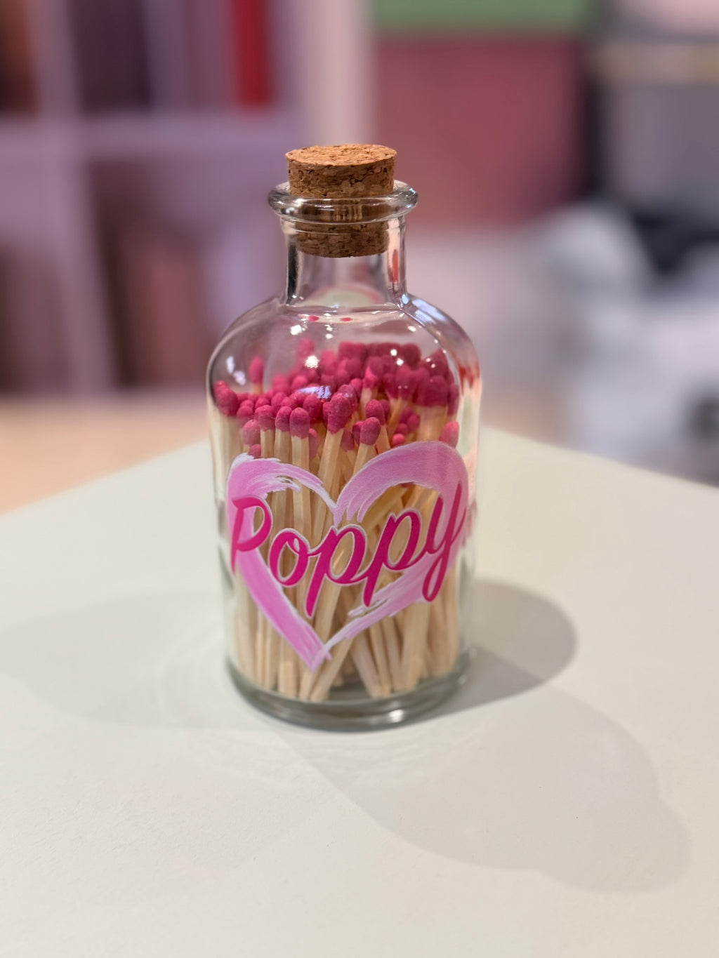 Clear glass bottle matches with cork lid filled with pink matches and 'Poppy' written on a white surface.