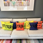 Colourful personalised silk pillows with 'YAIR' text on a beige sofa against a grey wall.