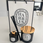 Two black candles with wooden lids and Diptyque-style branding in front of a Diptyque-style branded box.