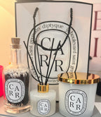 Diptyque style dffuser, candle, and match bottle with decorative sticks against a Diptyque-style  branded background.