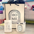 Gift bag, candle, and small box with 'HOME' and 'Shabbat at the Levines' text on a blurred background.