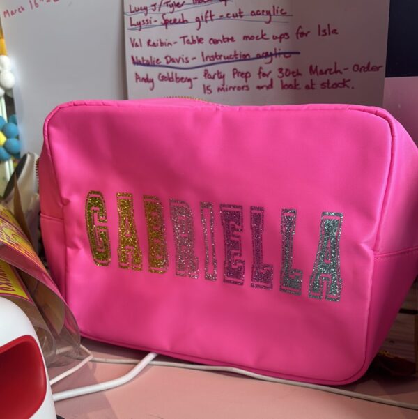 Pink washbag with 'GABRIELLA' in glittery letters on a desk with papers in the background