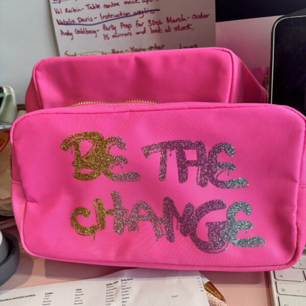 Pink washbag with 'Be the Change' text on a desk with papers and a computer.