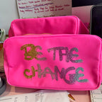 Pink washbag with 'Be the Change' text on a desk with papers and a computer.