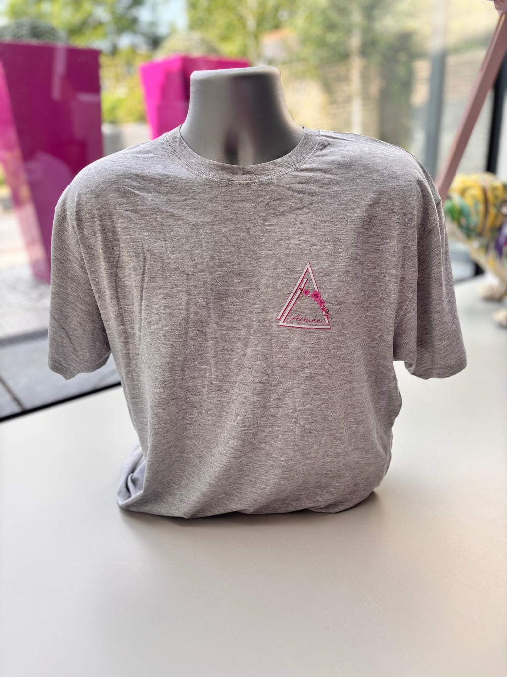 Grey event t-shirt with a pink triangle logo on a mannequin against a blurred indoor background
