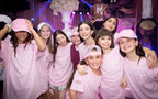 Group of young girls in pink event t-shirts posing together in a festive setting with purple lighting.