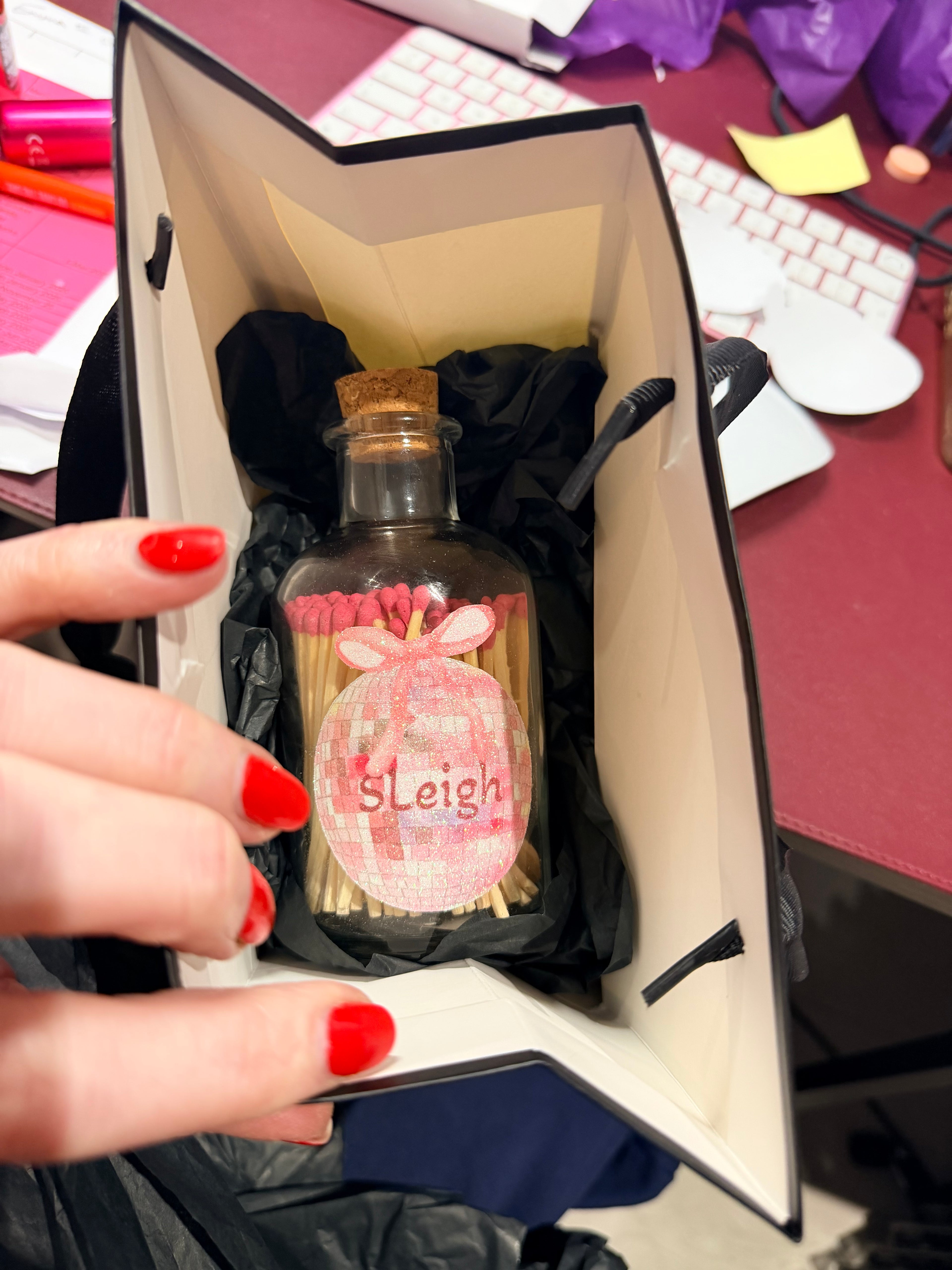 Hand holding a small match bottle with a pink label inside an open box on a desk.