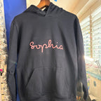 Black hoodie with 'Sophia' printed in pink on a hanger against a store background.