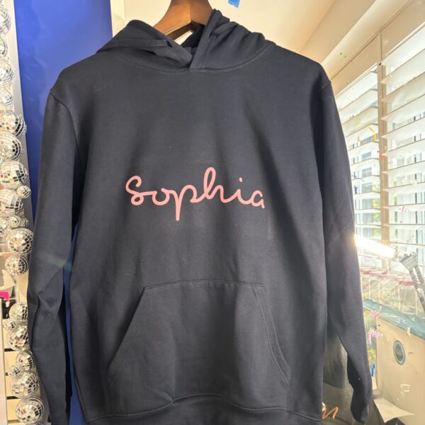 Black hoodie with 'Sophia' printed in pink on a hanger against a store background.