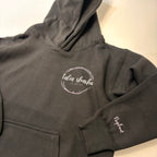 Dark grey hoodie with a logo on the chest and text on the sleeve against a light background