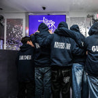 Group of people wearing hoodies with names on the back, gathered around a screen displaying a logo.
