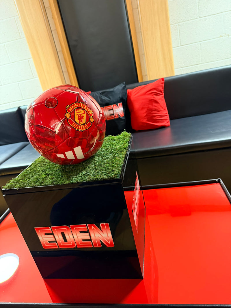 Red football labelled 'Manchester United' on a black stand with 'EDEN' written on it, placed on grass as a gift card box
