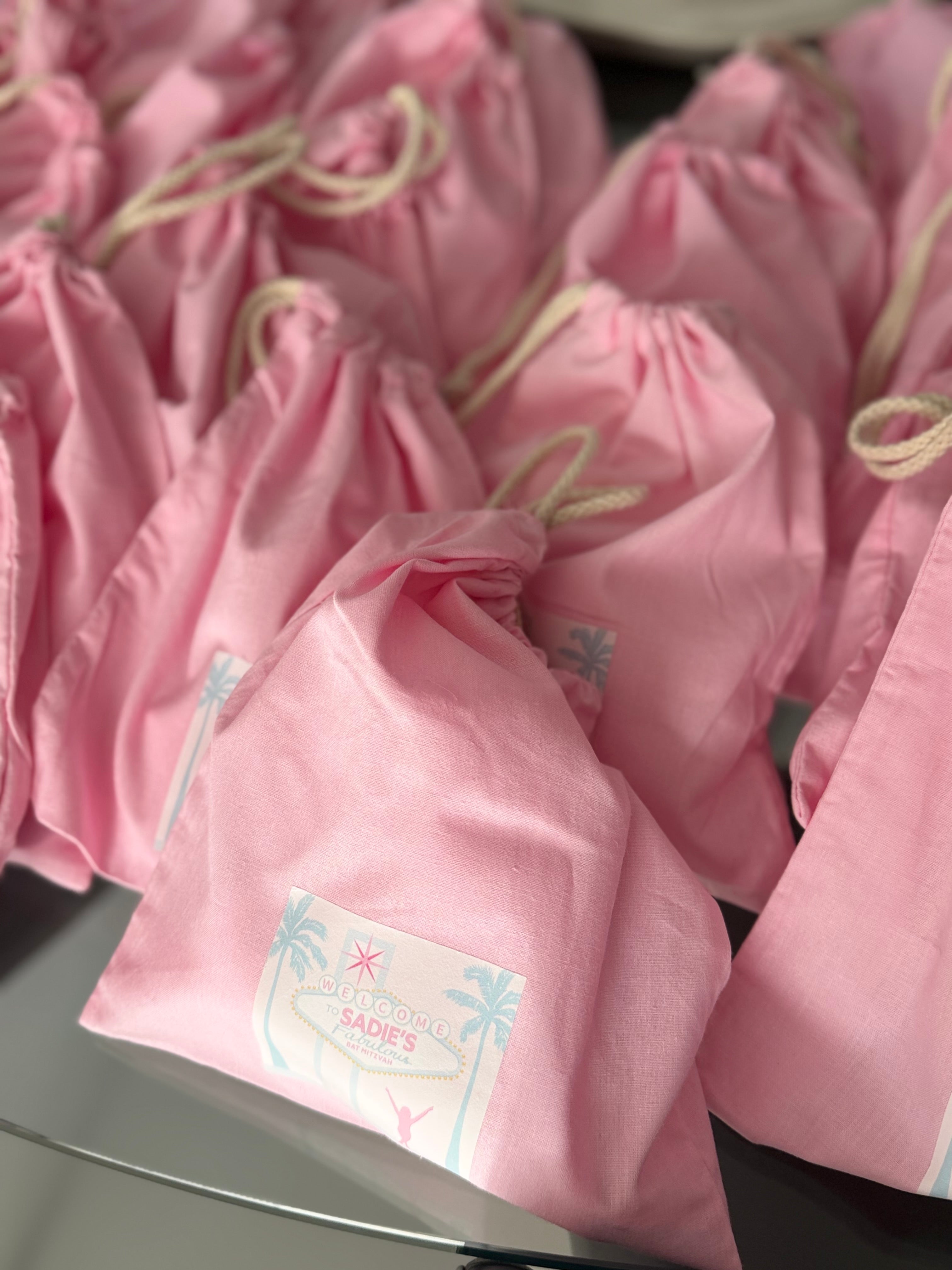 Pink drawstring personalised tote bags with labels on a reflective surface