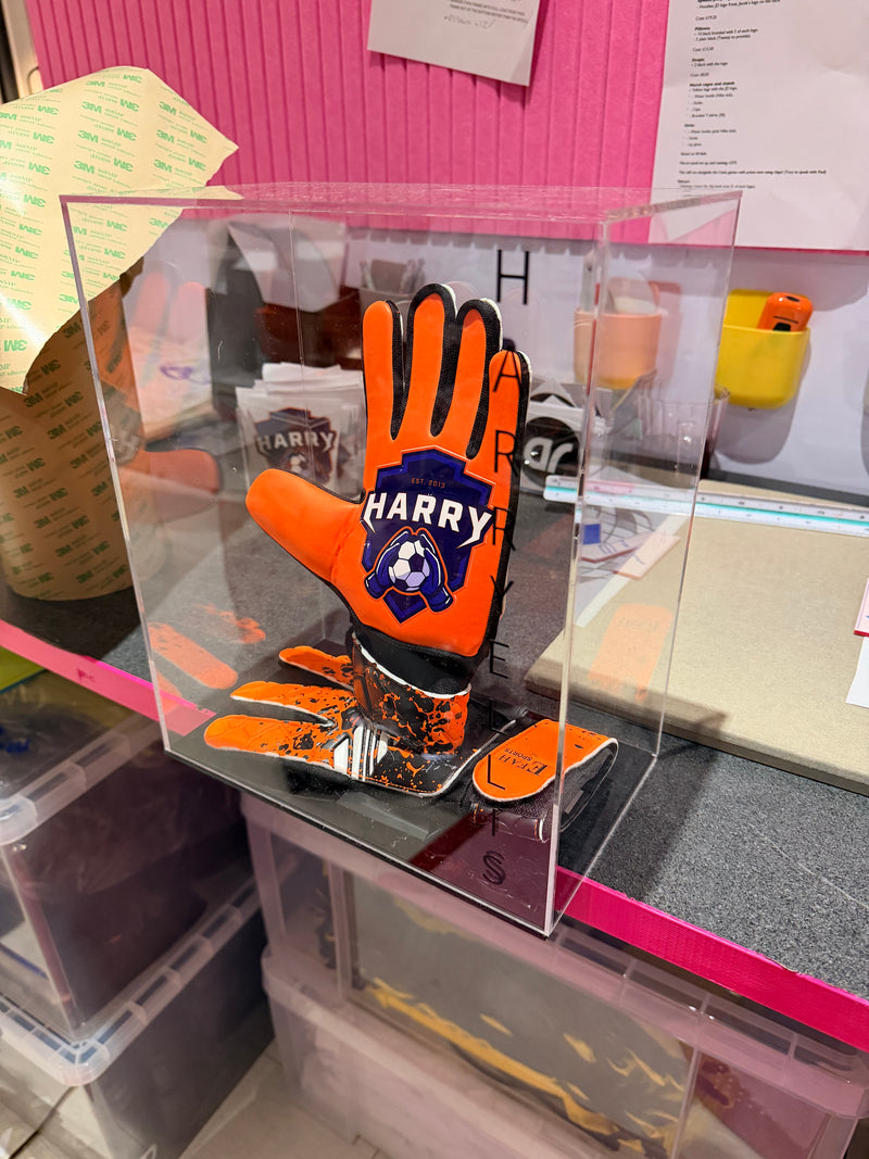 Personalised orange football goalie gloves with a logo on a clear stand against a cluttered background