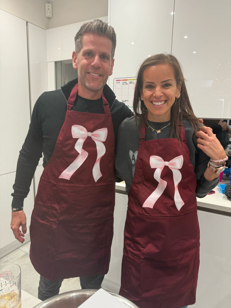 Two people wearing personalised maroon aprons with pink bows in a kitchen.