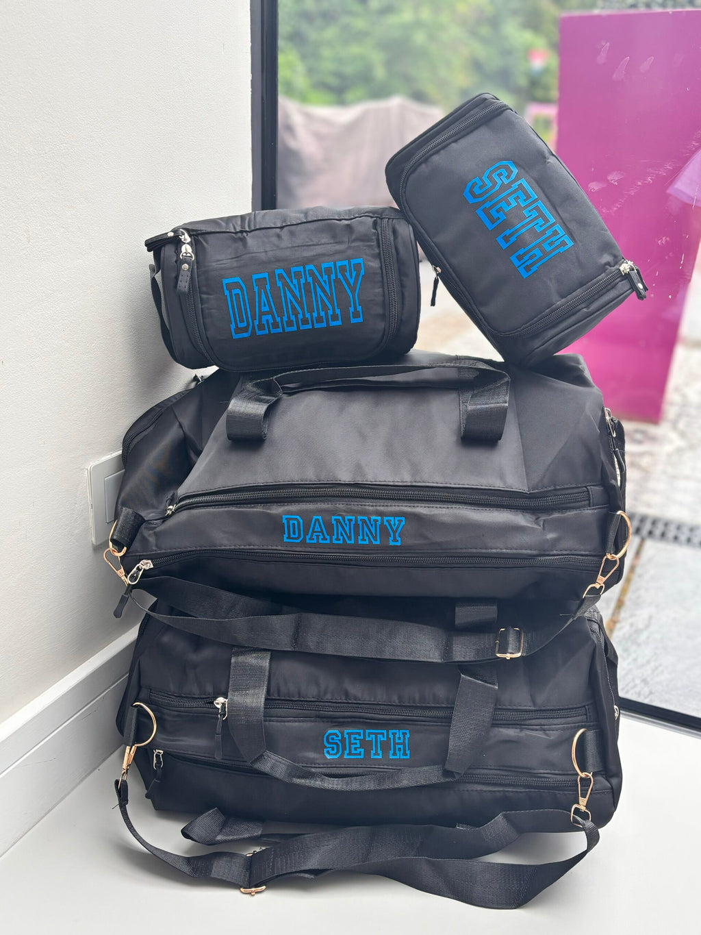 Black washbags and duffel bags with names embroidered on them, including 'DANNE' and 'SETH', against a blurred indoor background.