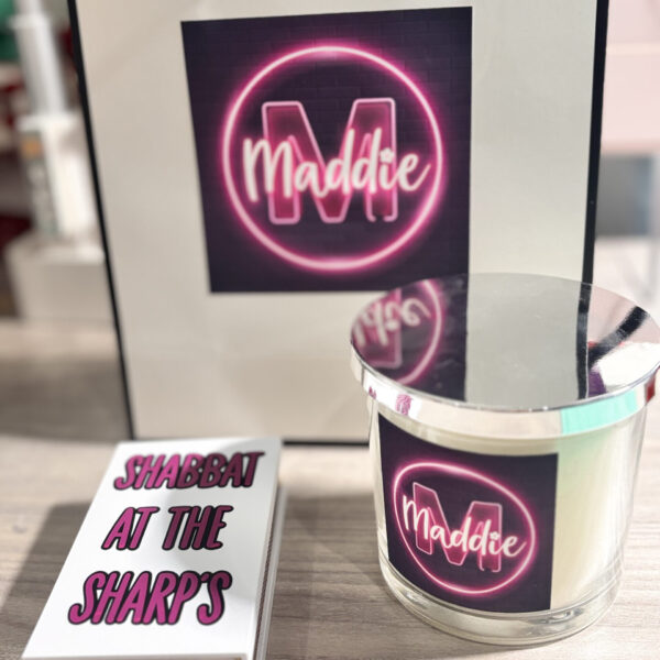 White match box sleeve with 'Shabbat at the Sharps' text, with a candle labelled with a neon design with 'Maddie' on a blurred background.