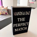 Black match box sleeve with 'The Perfect Match' by MANDS & DAZ on a table in a room.