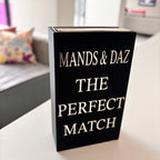 Black match box sleeve with 'The Perfect Match' by MANDS & DAZ on a table in a room.
