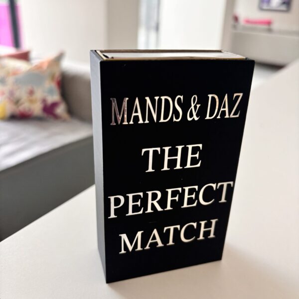 Black match box sleeve with 'The Perfect Match' by MANDS & DAZ on a table in a room.