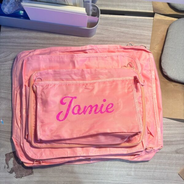 Pink packing cubes with 'Jamie' embroidered on it on a wooden surface