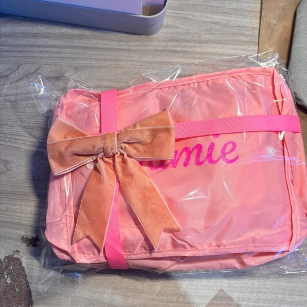 Pink packing cubes with a brown bow and 'Amie' written on it, wrapped in clear plastic on a wooden surface.