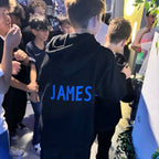 Person wearing a black hoodie with 'JAMES' printed on the back, surrounded by people.
