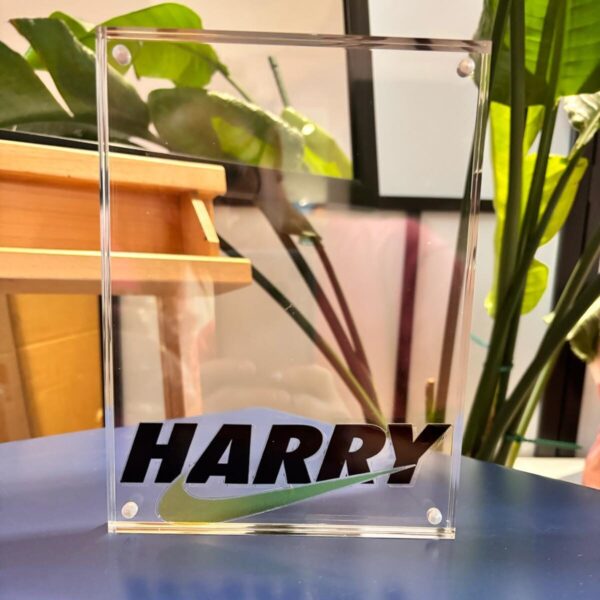 Clear acrylic photo frame with 'HARRY' engraved on it, placed on a reflective surface with plants in the background.