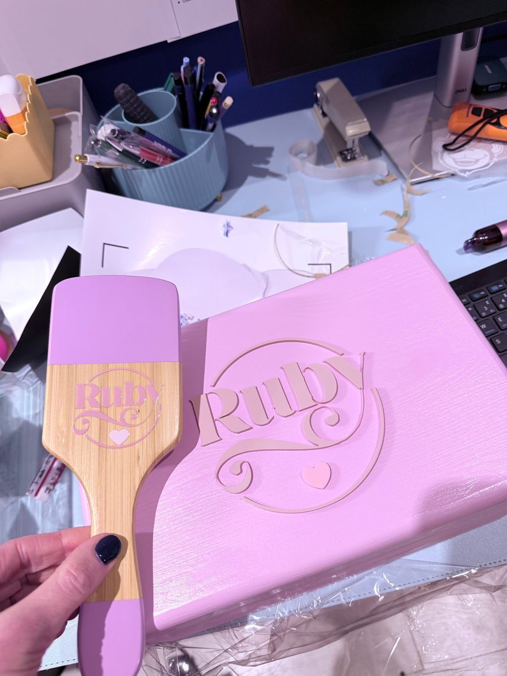 Pink keepsake box and matching pink and wooden brush with 'Ruby' branding held over a desk.