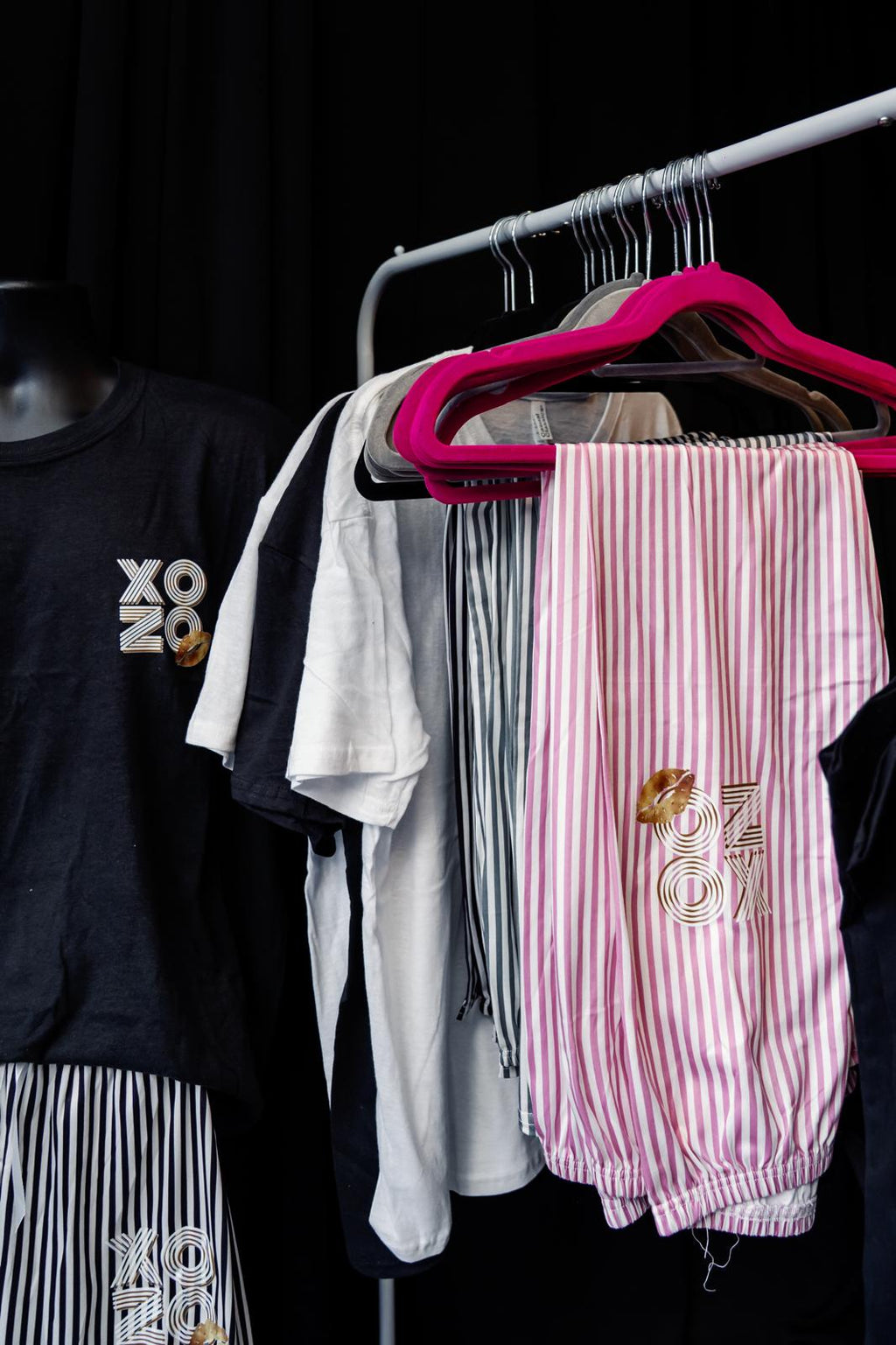 Pyjama clothing items including a black t-shirt with 'XO' logo, white t-shirt, pink striped pants, and a black jacket on a rack.