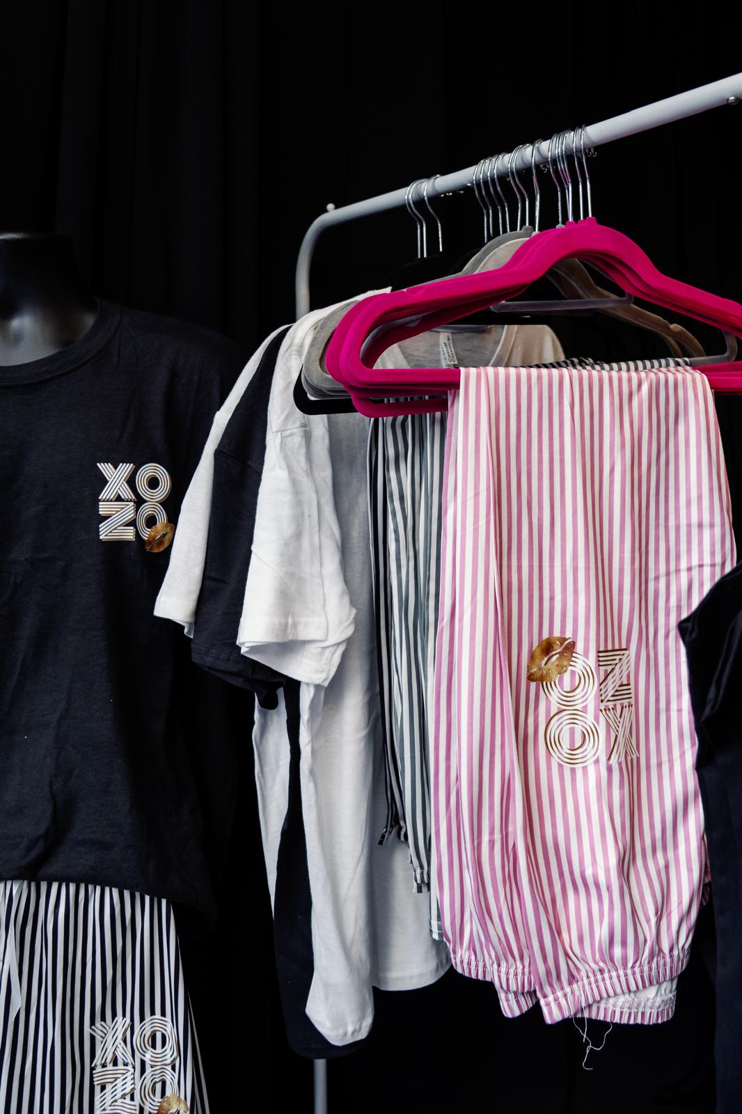 Pyjama clothing items including a black t-shirt with 'XO' logo, white t-shirt, pink striped pants, and a black jacket on a rack.
